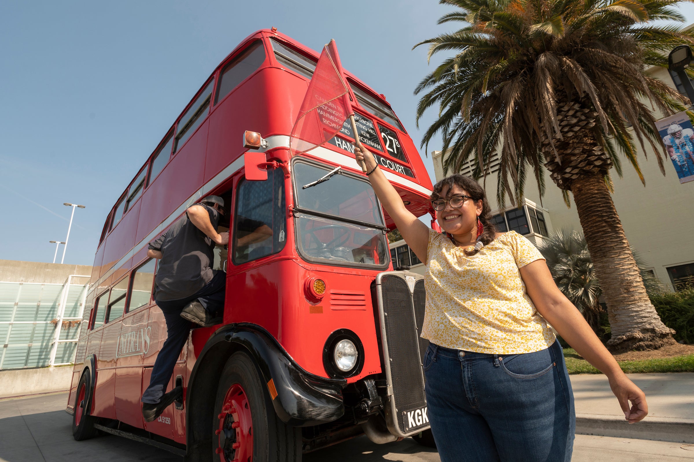 A student stands in front of a double-decker bus waving a red flag at UC Davis. 