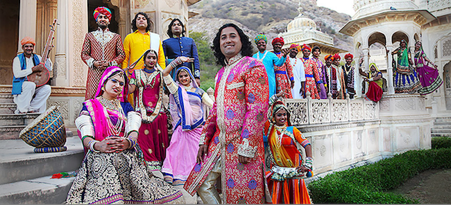 The band members standing in front of an Indian builing, wearing brightly colored costumes.