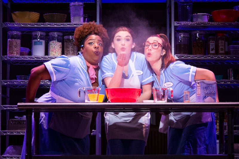 Three waitresses standing at a table, making a pie and blowing a handful of flour in the air.