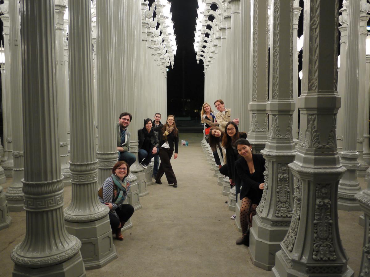 A class of students peaks out from behind white columns in a building. 