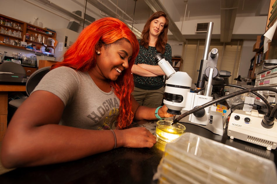 A student smiles as she inspects yellow liquid underneath a microscope, while her professor watches on. 