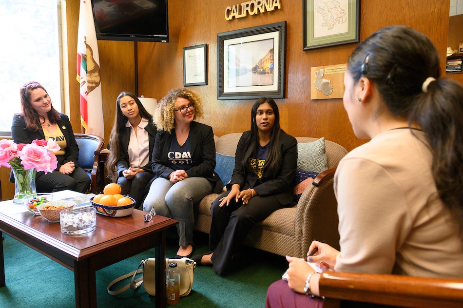 Students in UC Davis T-shirts and blazers talk around couches in an office. 