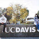 Students wearing face coverings sit on UC Davis sign