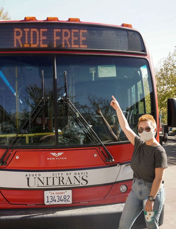 Female bus driver stands in front of bus, pointing to "Ride Free" electronic sign.