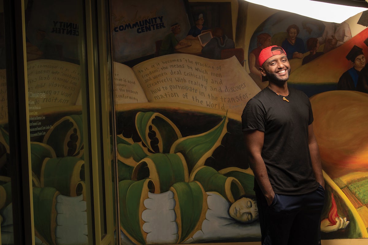 A smiling sociology major in a red baseball cap stands in front of a colorful mural. 