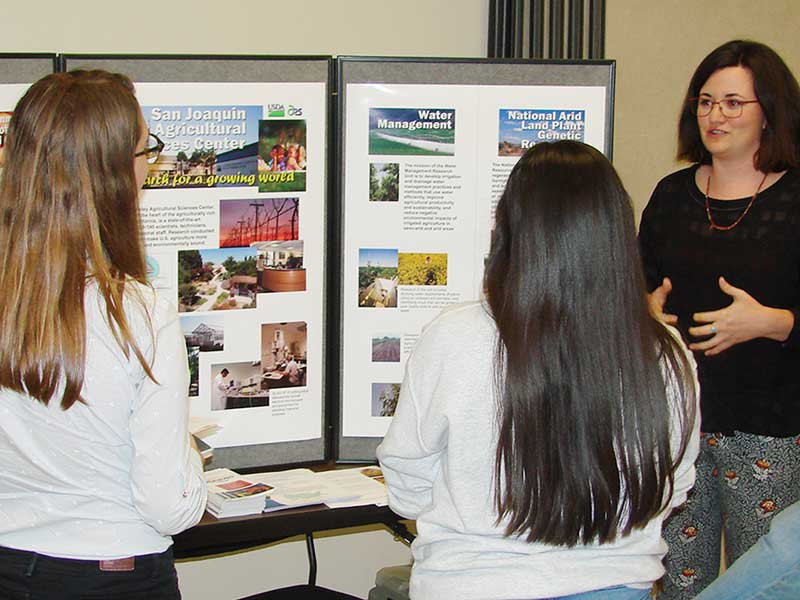 Woman on right addresses students with presentation boards in the background