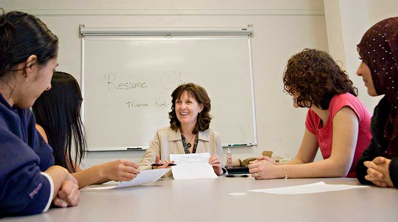 Woman at center of conference table with female students on either side listening to her