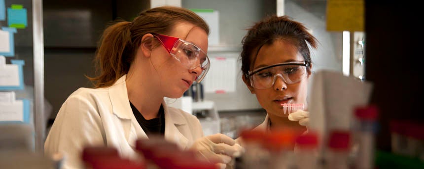 Two young women in lab coats look closely at a test tube