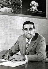 Black and white photo of a white man with dark hair wearing a jacket and tie sitting at a desk. Above and behind is head is a photo of a mushroom cloud. 