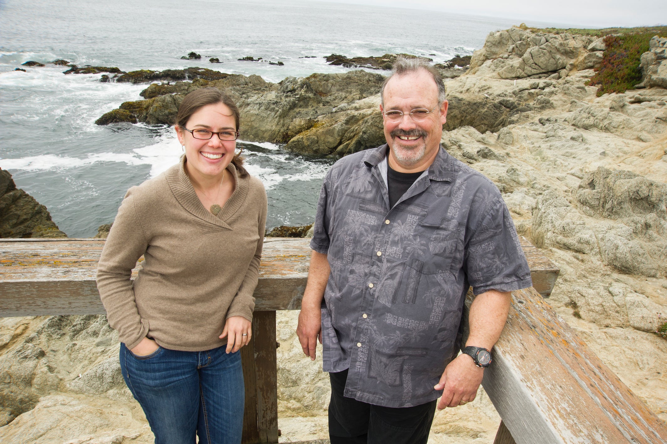 Female and male scientists face camera smiling on a deck with ocean in background