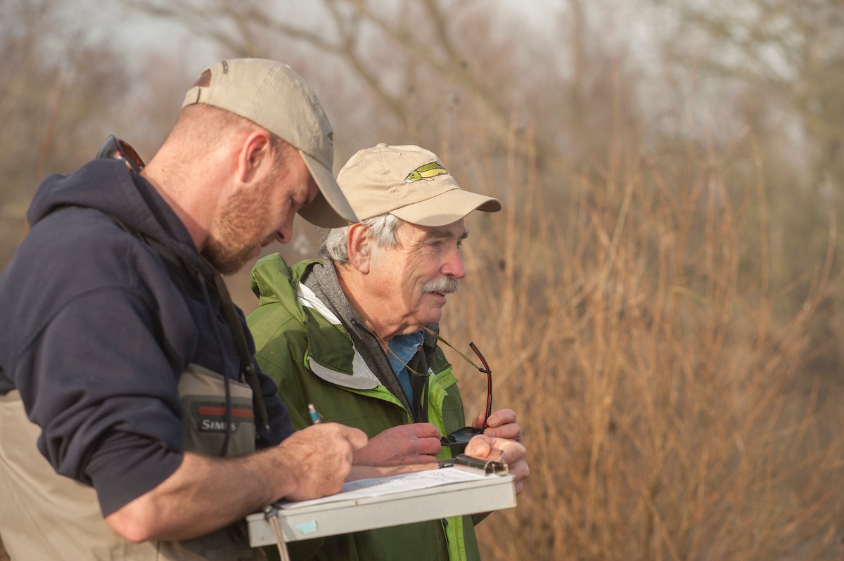 Two men in tan ball caps take notes outside with brown trees in background