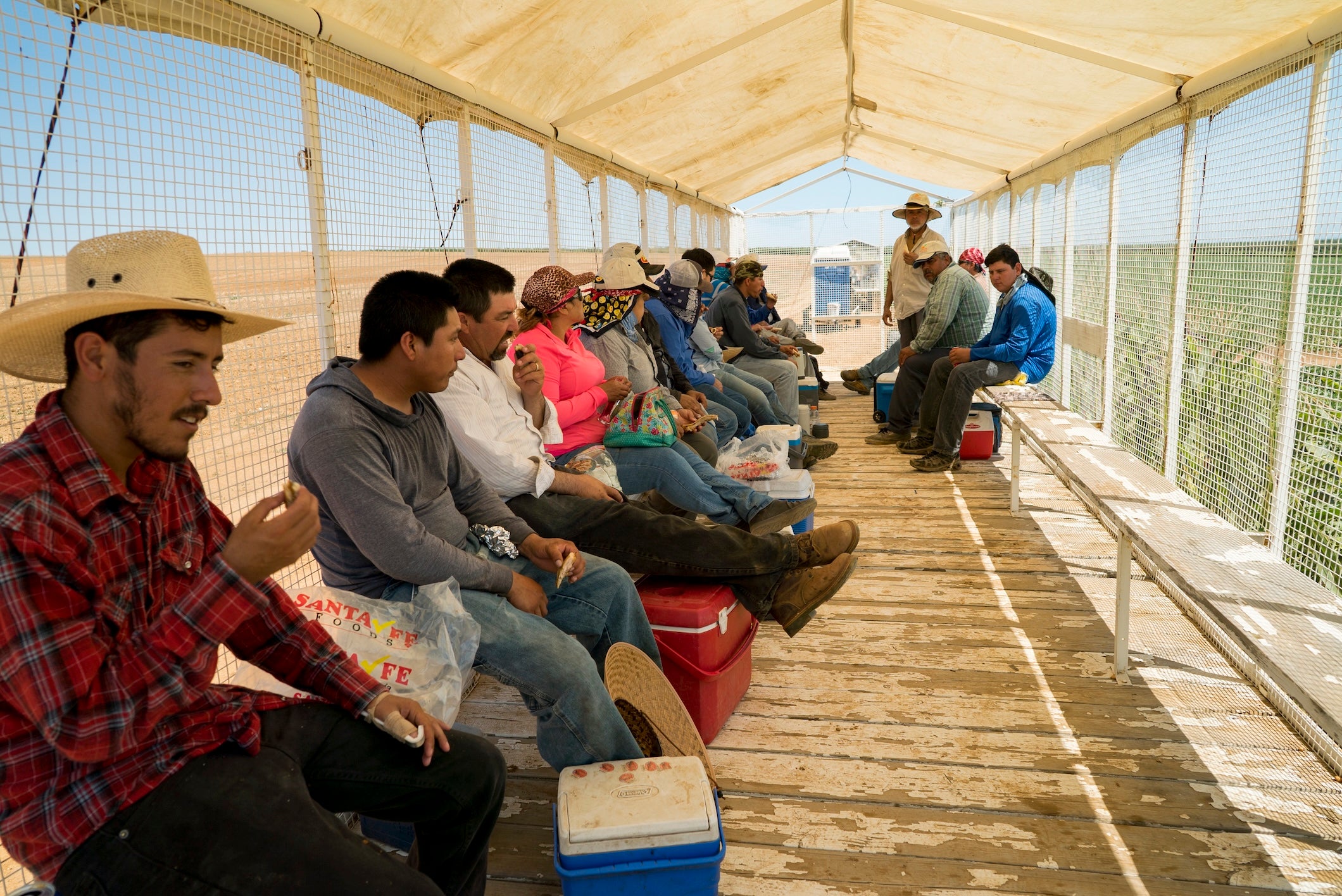 Workers seated along benches inside an open-sided covered farm trailer, holding lunches and coolers