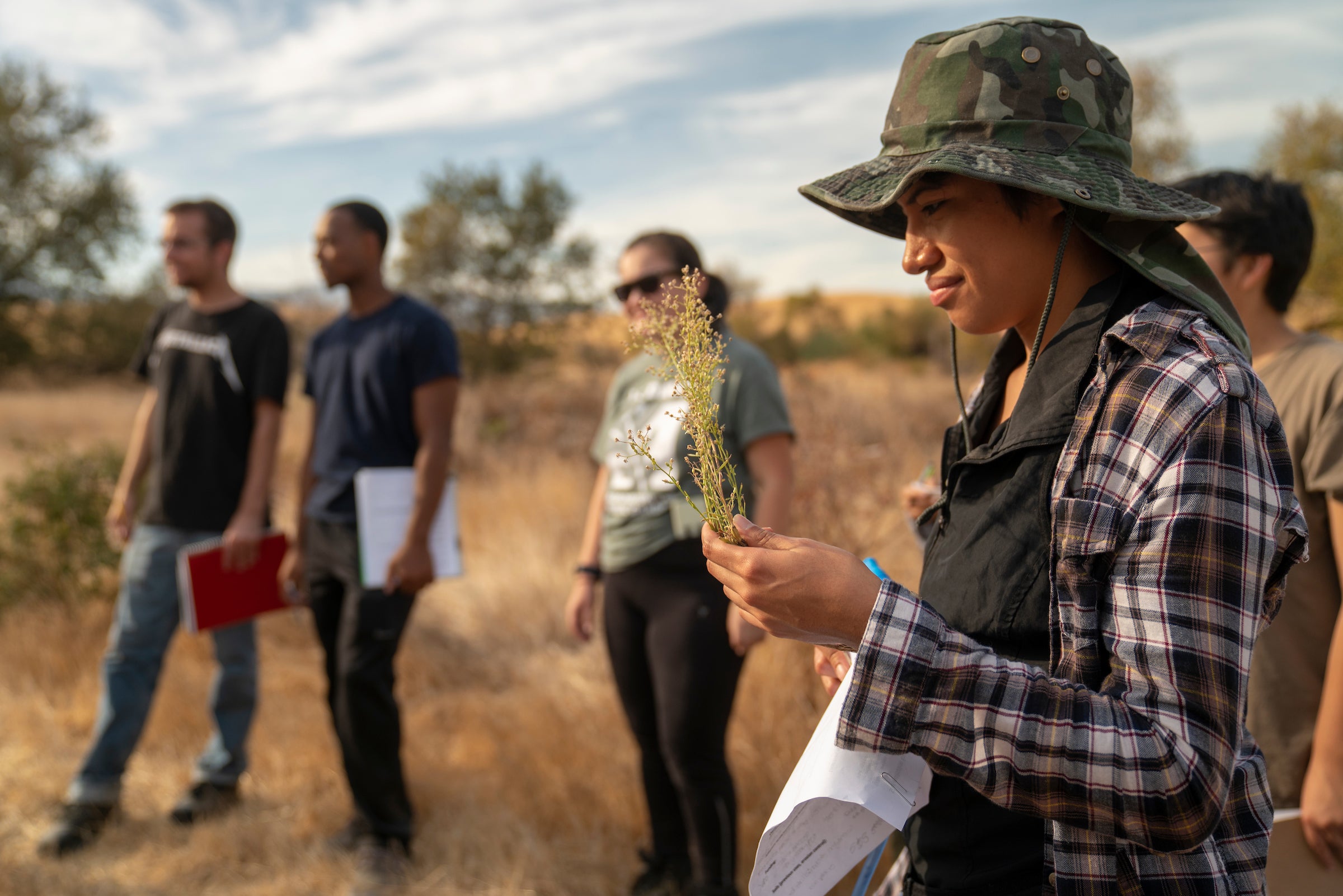 Woman in hat looks at stalk of dried plant in hand outdoors at ranch with three people in background