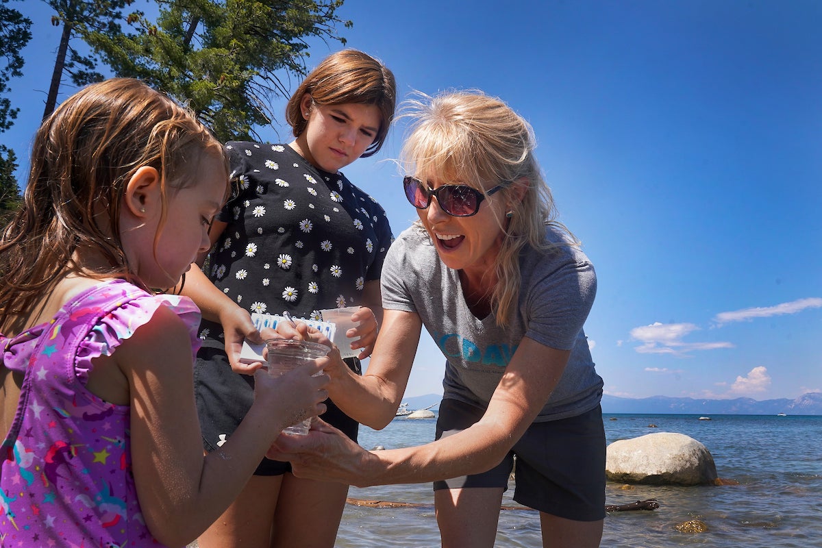 Woman in sunglasses and two girls at a lakeshore, smiling as they examine a small marine creature