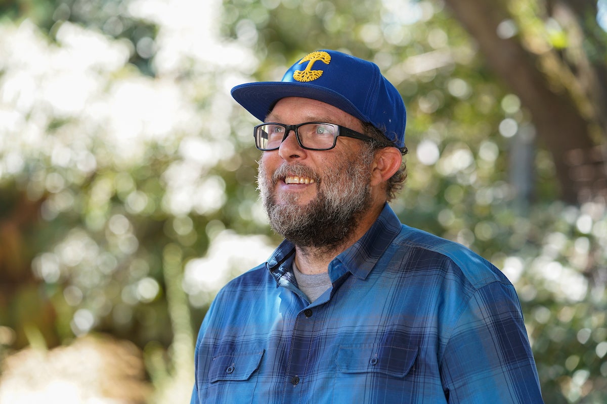 Man in plaid shirt, glasses and ball cap smiles looking in distance with trees in background