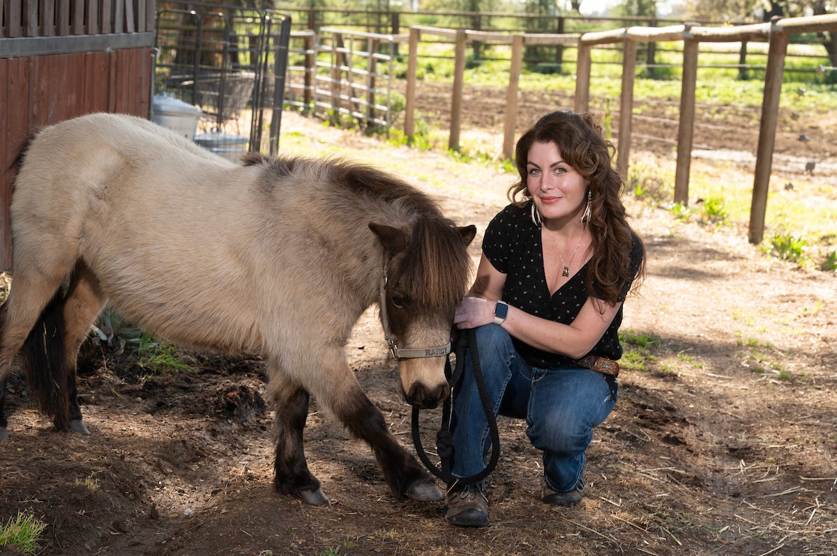 Woman kneeling and smiling next to a small tan pony in a sunlit fenced paddock