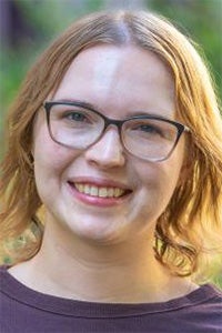 Outdoor headshot photo of smiling woman with glasses and shoulder-length blonde hair