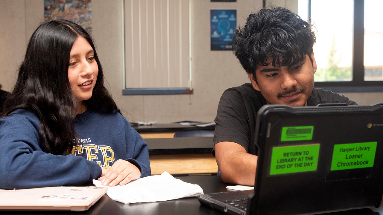 A woman sits at a table with a student working at a laptop.