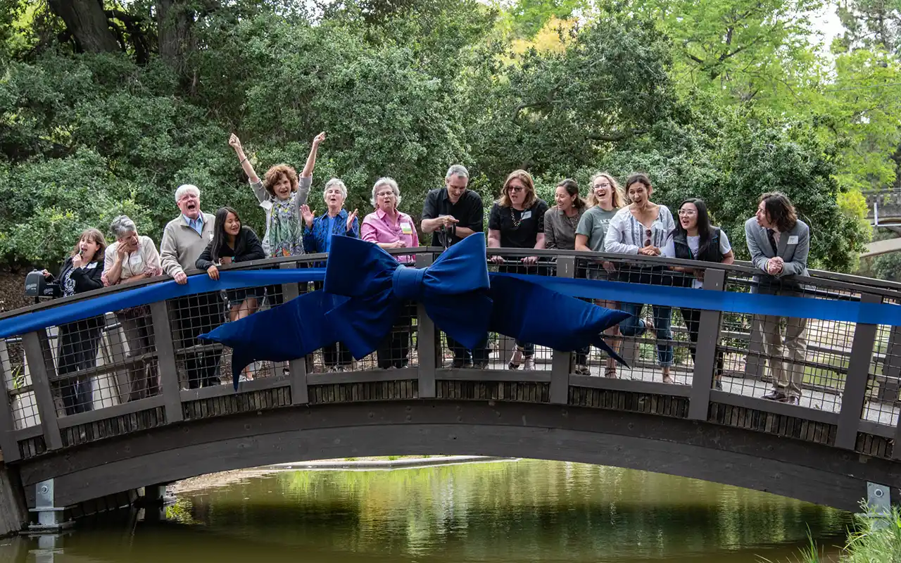 Cheering group on wooden arched bridge, while one holds large scissors to cut a blue ribbon stretched across the span of the bridge