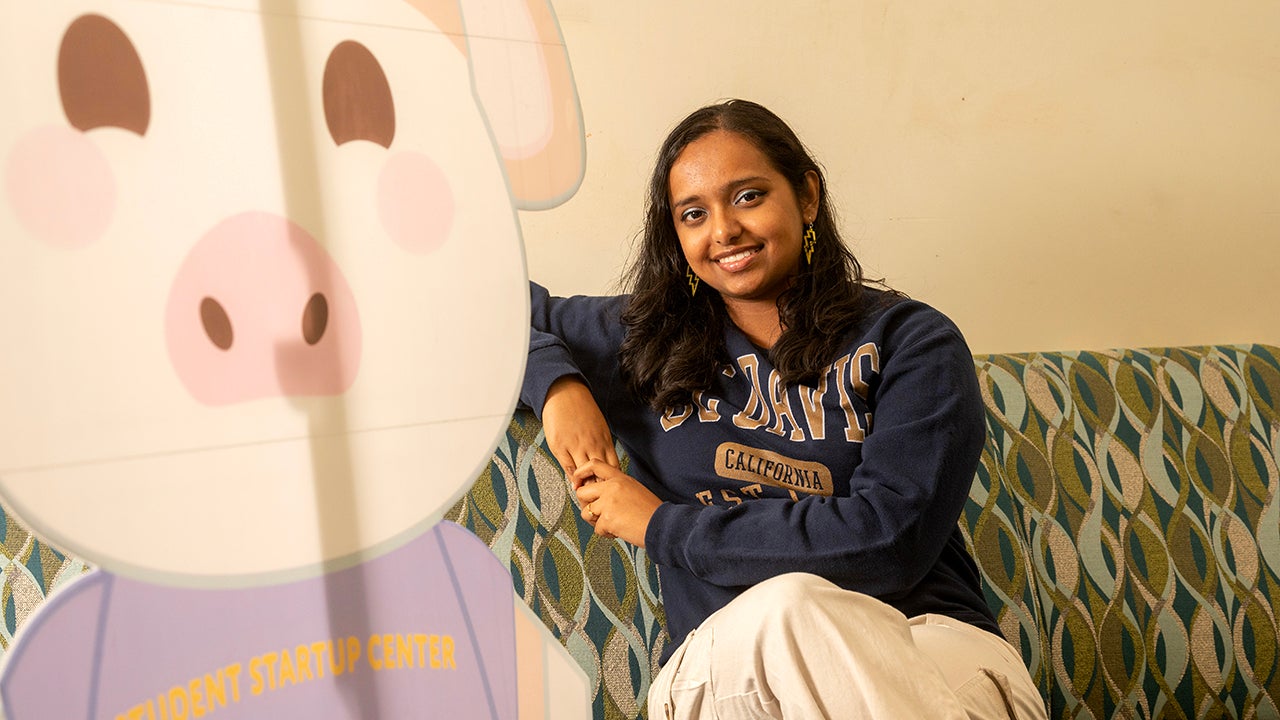 Young woman in navy UC Davis sweatshirt sitting on a sofa, beside a large cardboard cut-out of the Student Startup Center's mascot.