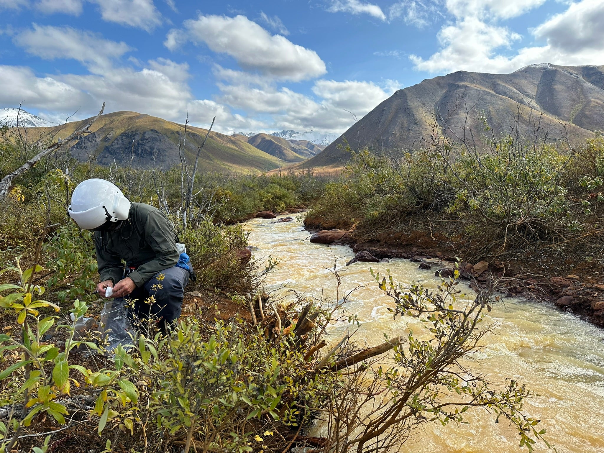 Scientist in protective white helmet crouching by muddy stream in Alaska, mountains under blue sky