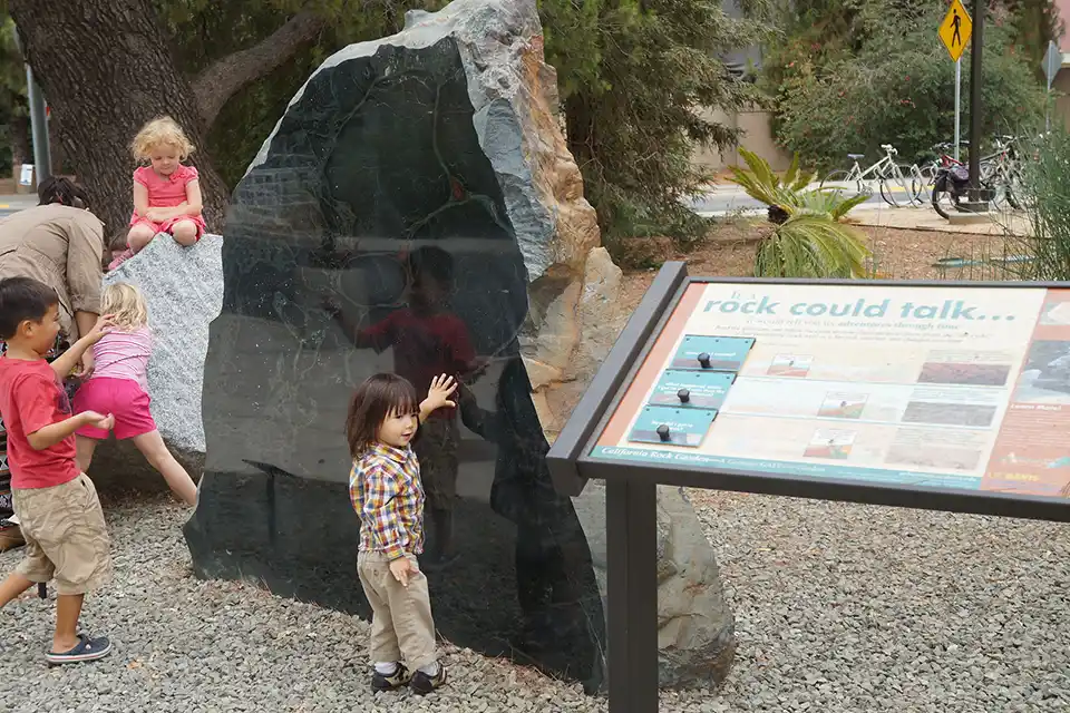 Children examining a polished dark rock next to an outdoor interpretive sign