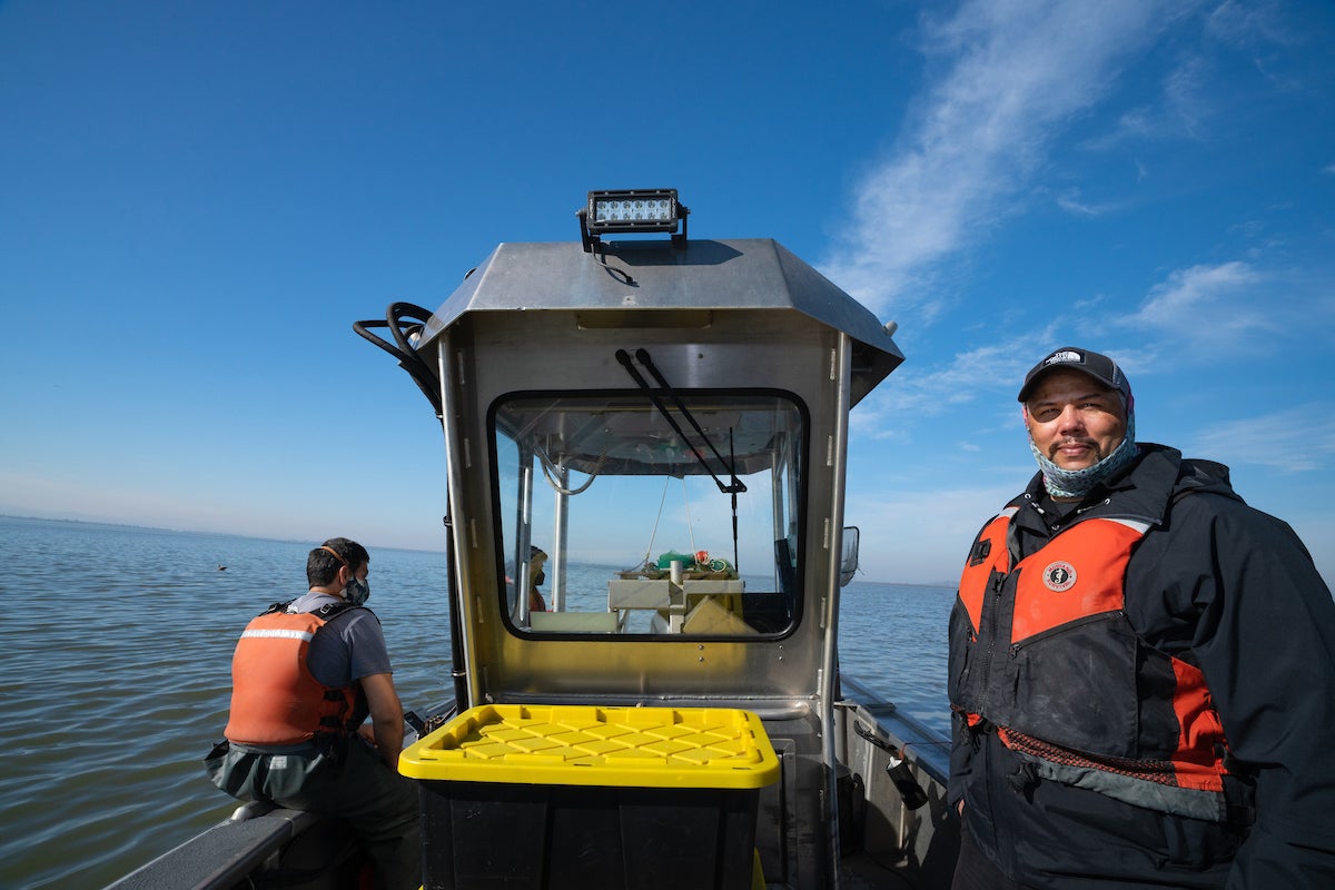 Levi Lewis look at camera aboard research vessel while other researcher looks at bay