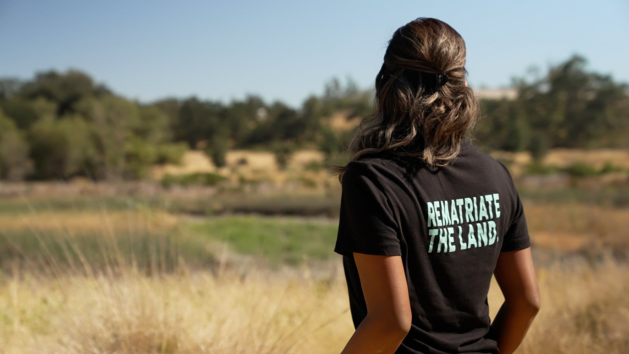 Back of Native American woman looking toward field. Back of her shirt says "Rematriate the Land."