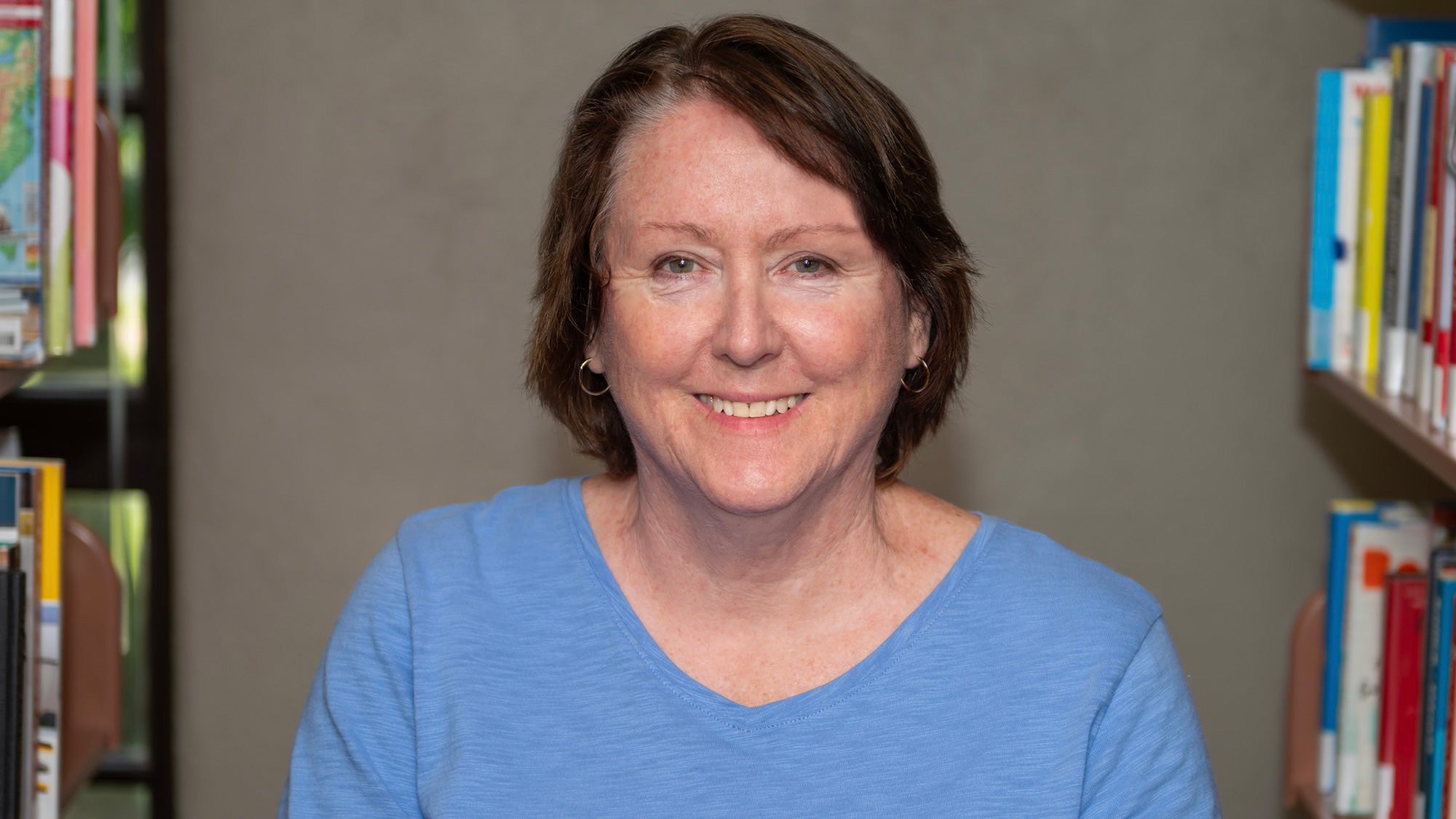 Smiling woman with short brown hair, wearing a blue sweater, in a library setting.