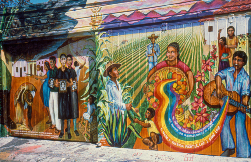Large mural showing farmers in front of rich fields, smiling, guitar player and words from a song; on the left are a group of women holding black and white photo of murdered family with armed men to their left.