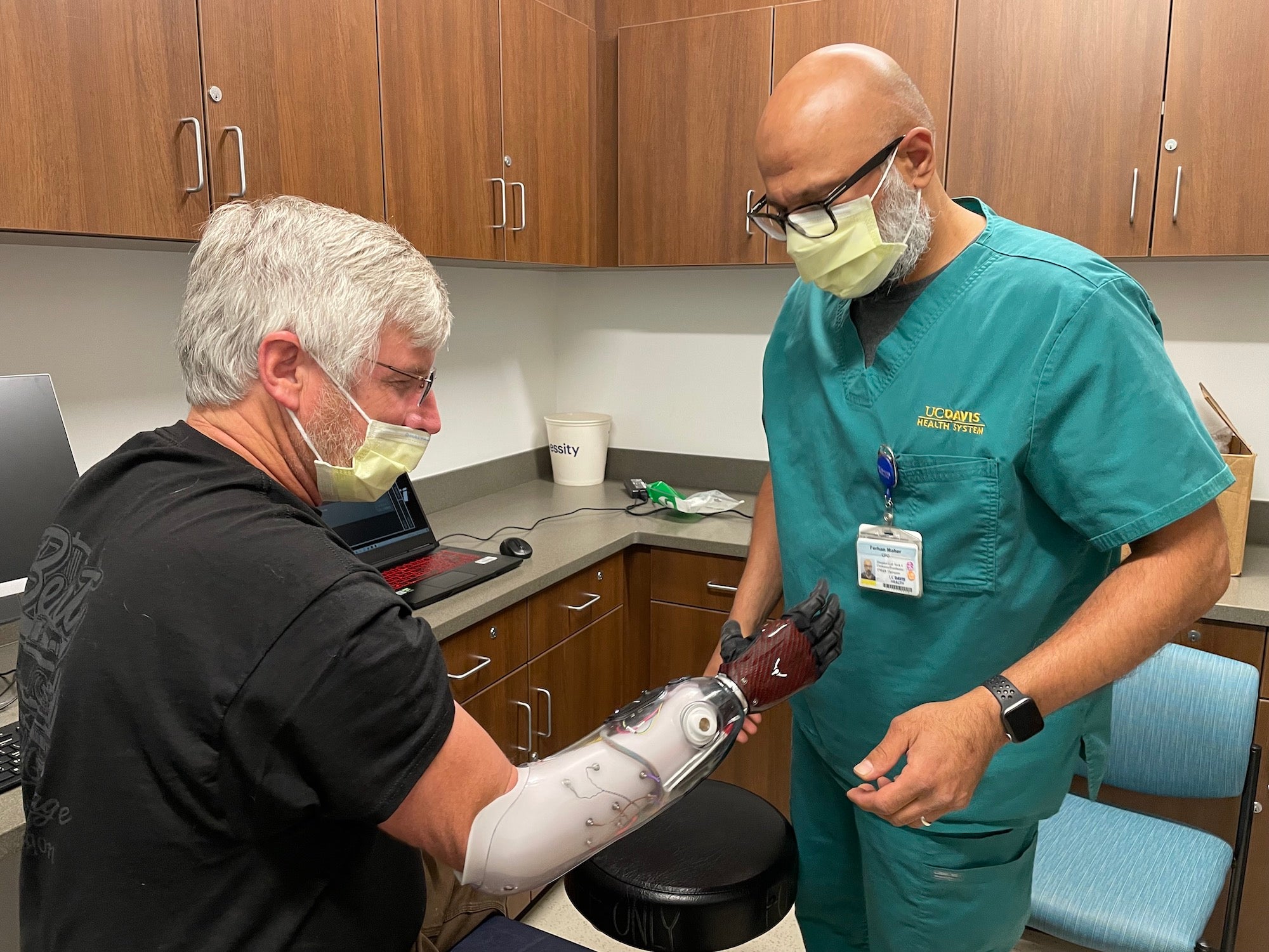 Dave Brockman, a hand amputee, wears a myoelectric prosthesis that looks like a red and black glove. He sits in front of his orthotist who is making sure the fit is correct.