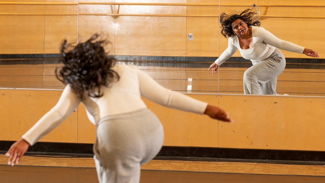 A woman in gray sweatpants and cream top dances facing a studio mirror.
