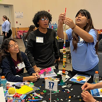 Three students at a workshop table collaboratively building a tall LEGO tower