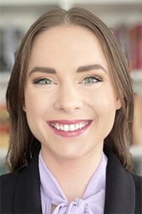 Headshot of smiling woman with short brown hair, lavender blouse and blazer, blurred bookshelves