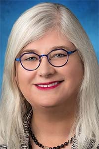 Professional headshot of smiling woman with long silver hair, blue-rimmed glasses, red lipstick