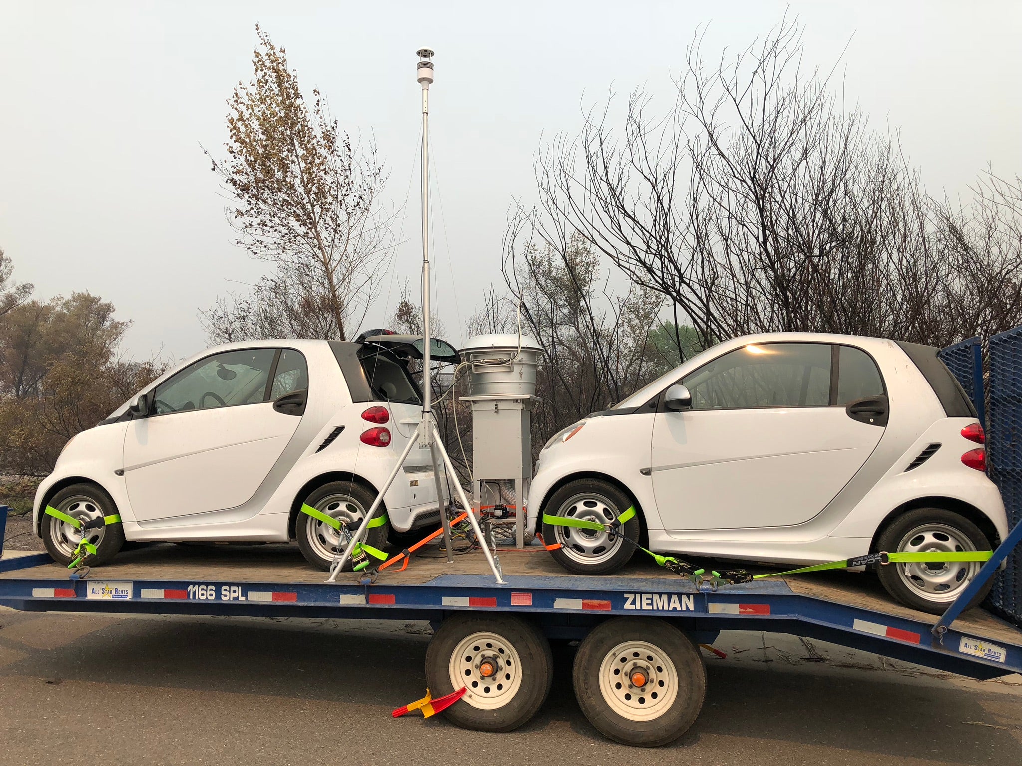 two white tiny cars on a trailer being pulled under gray smokey skies