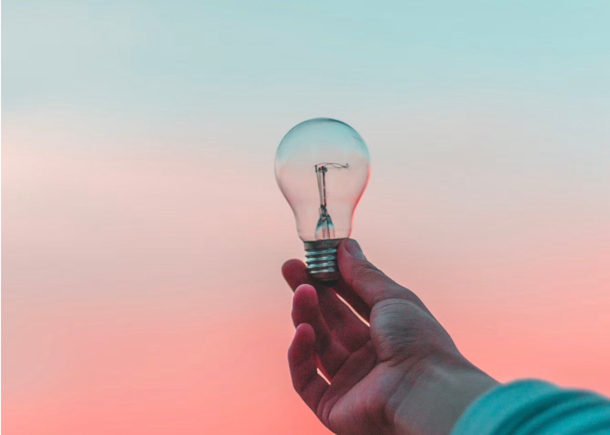 Hand holding clear light bulb against pastel blue-pink sky — photograph