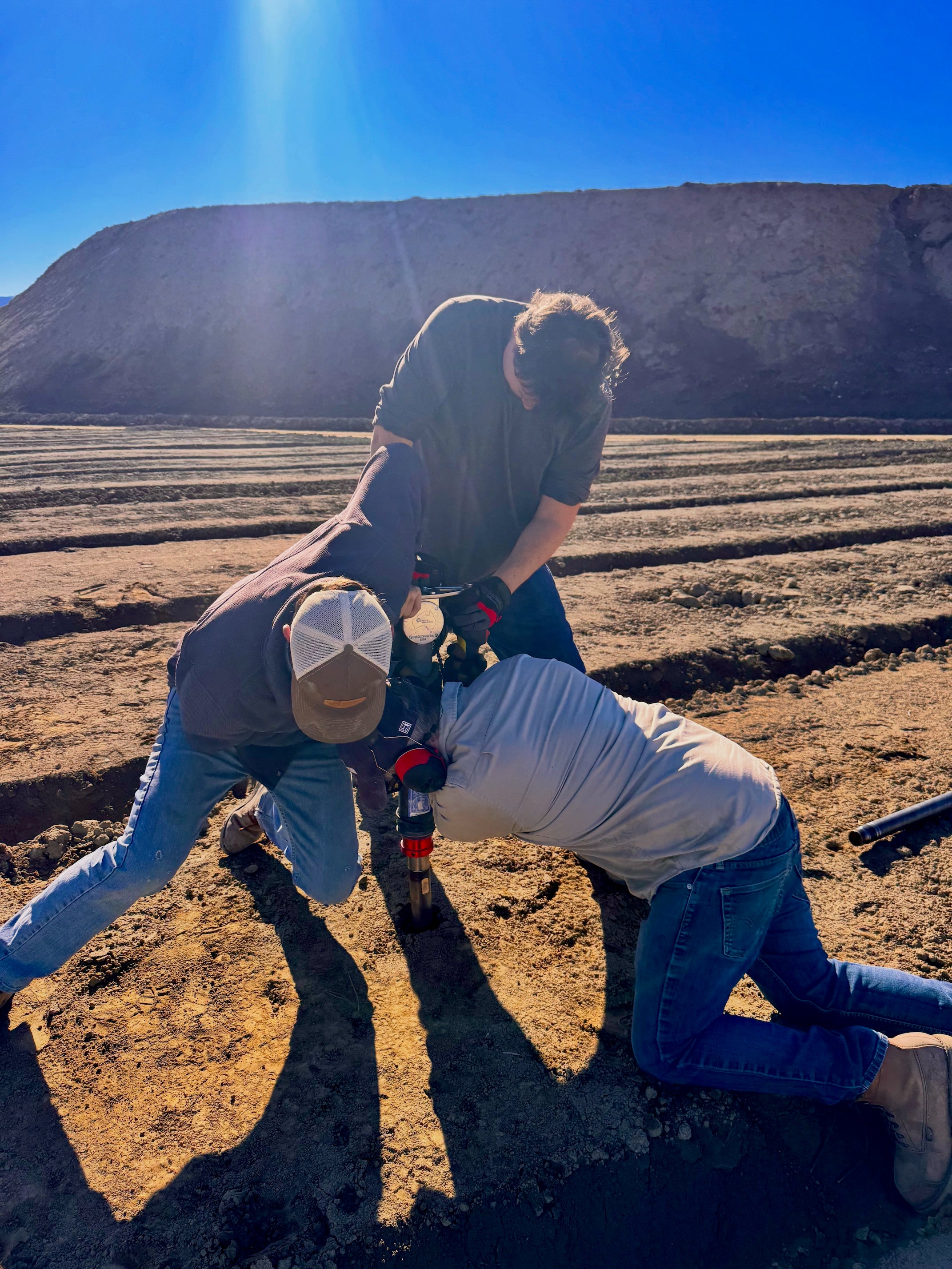 Three researchers bend over farm equipment, one kneeling, while exerting energy, on plowed field with mountain in background under blue sky