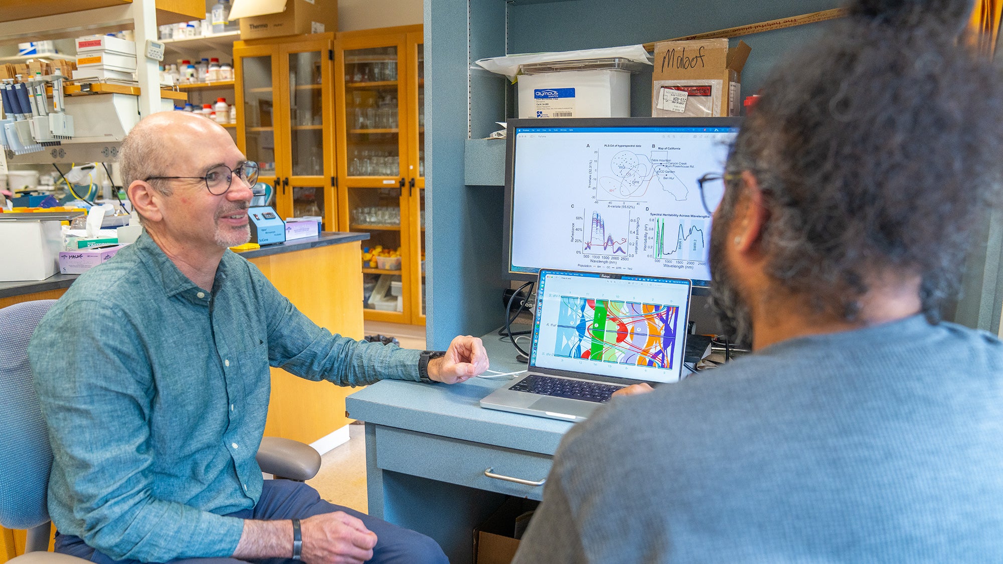 Male scientist wearing glasses points to colorful graphs on computer while colleague listens in lab