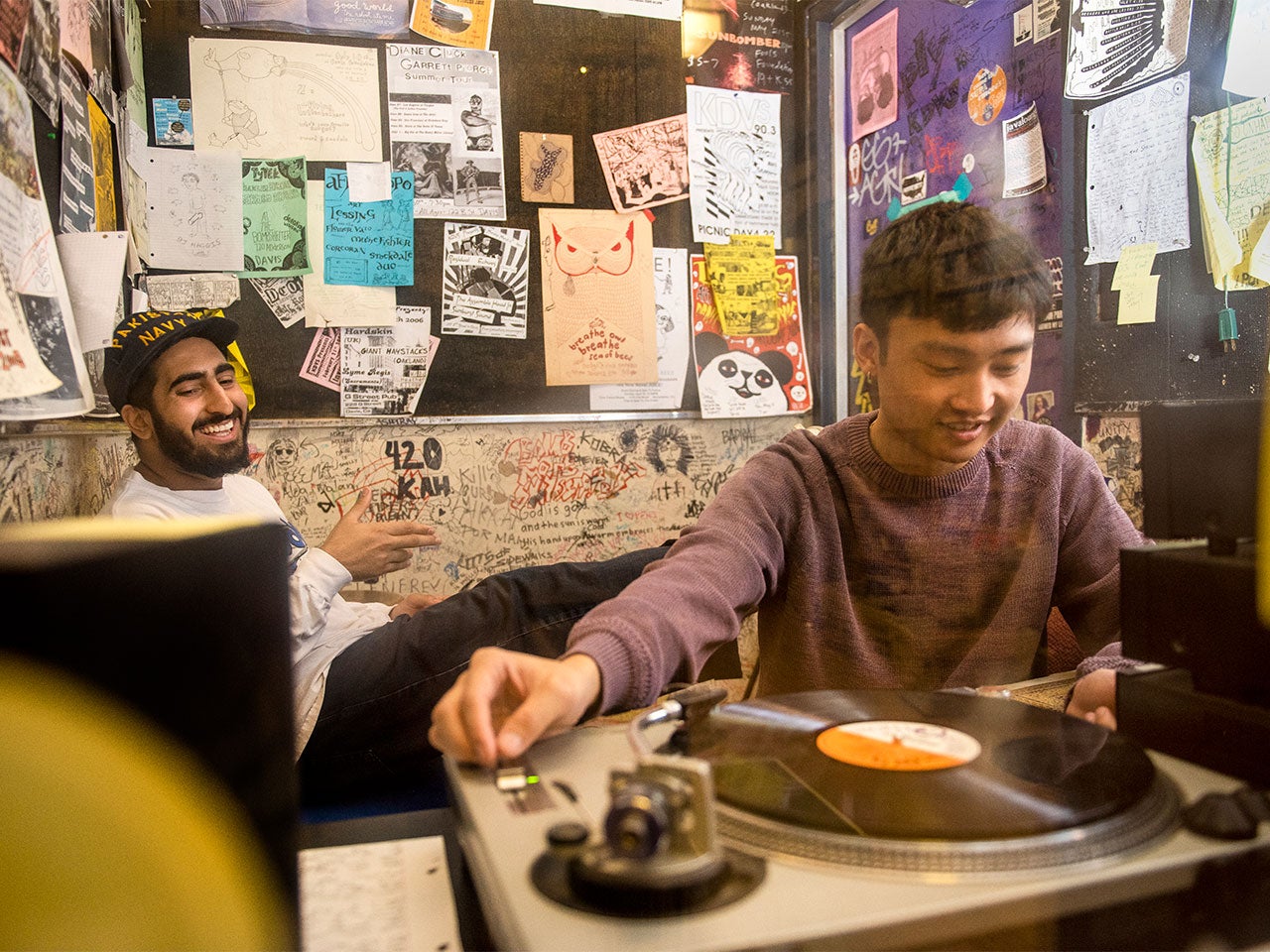 A UC Davis student works a record player in a radio studio while another student sits in front of the poster-filled back wall, smiling.