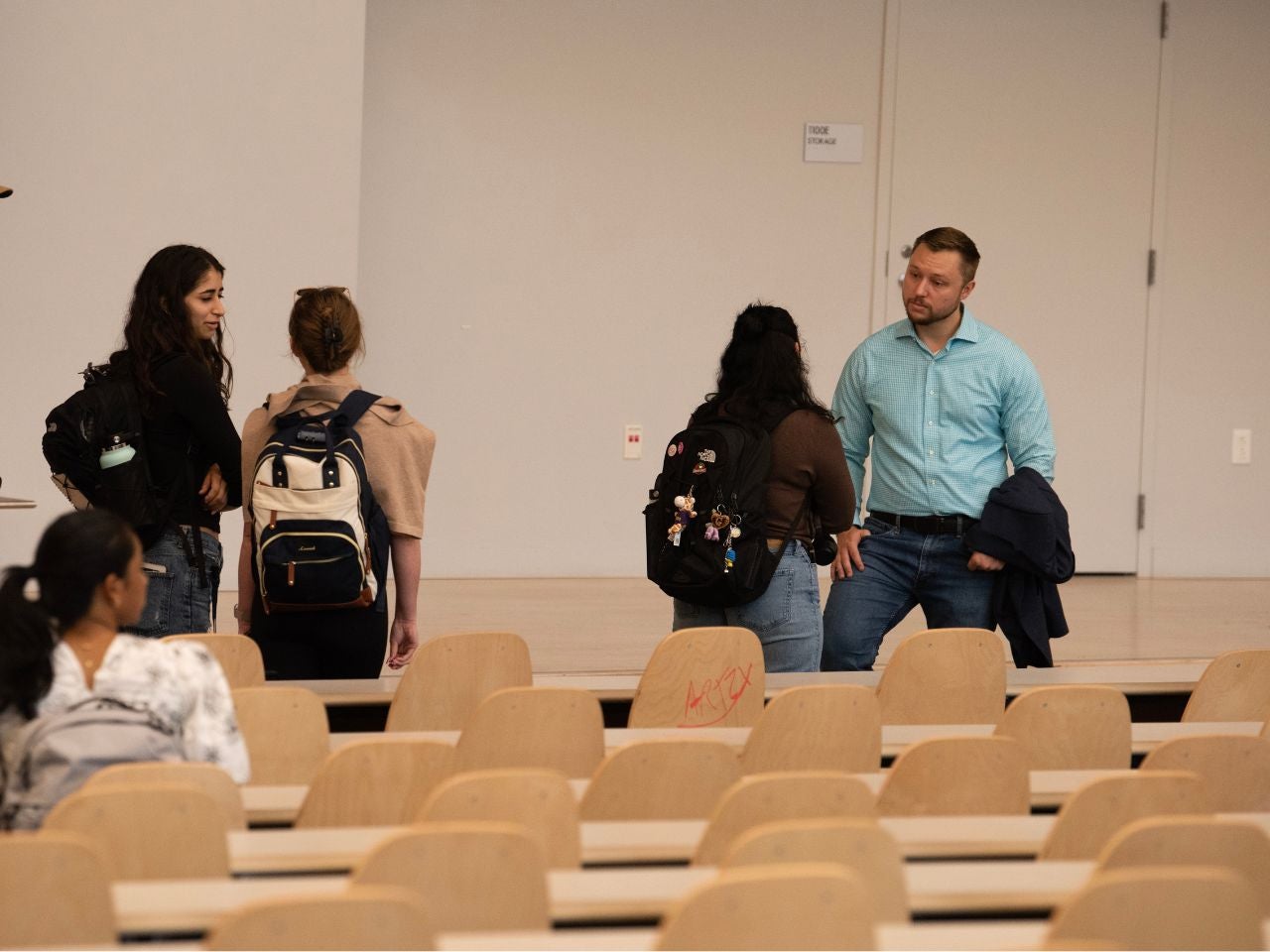 Faculty Lecturer Spencer Kiesel stands in conversation with several students at the front of a lecture hall.