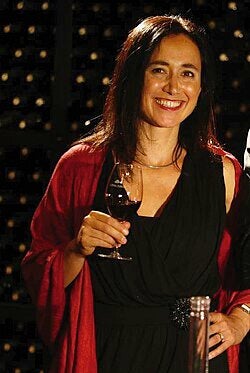 Portrait of a woman standing with a glass of wine, and appears to be in front of a rack of wine bottles