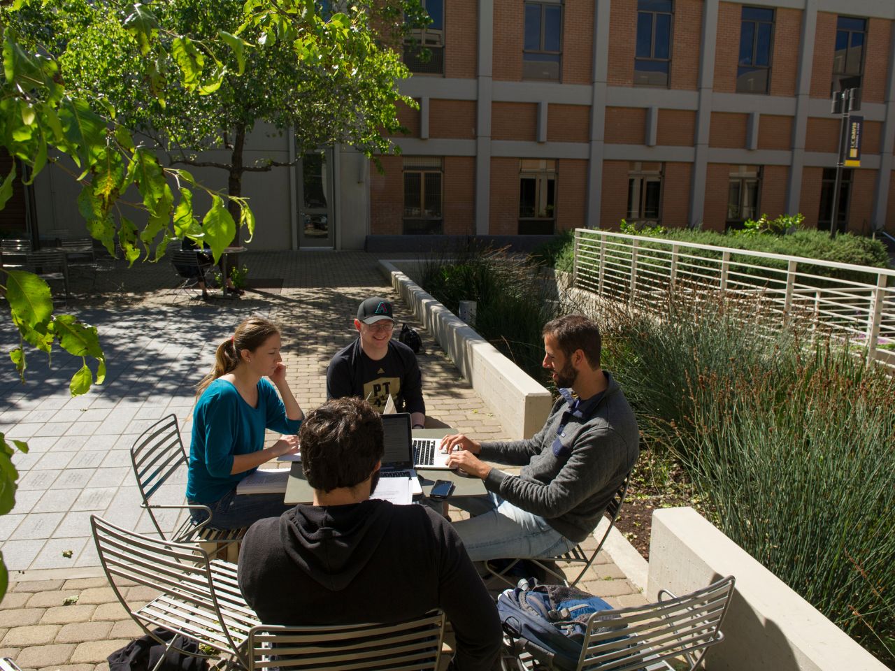 A group of students sit around a table in the courtyard of King Hall, talking and working on laptops.