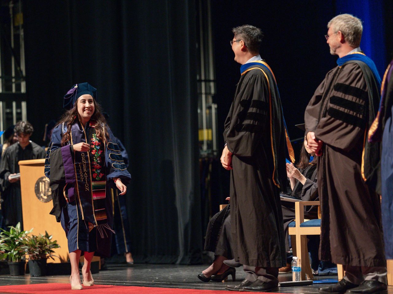 A student in a blue commencement gown and regalia, including a stole with the words "First Generation" walks across the stage at the UC Davis School of Law commencement ceremony.