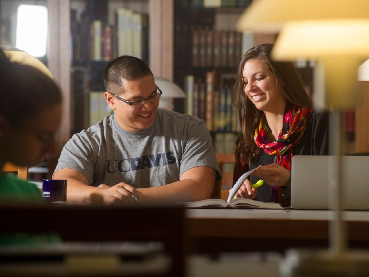 Two students sit at a desk in a library, smiling and looking down at the textbook open between them.