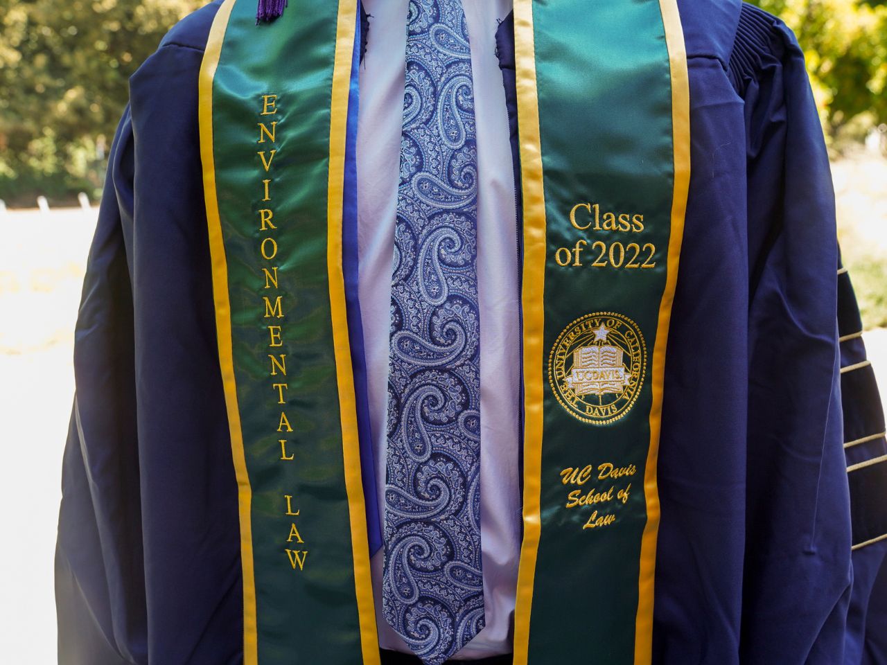 A student wearing a commencement robe and tie displays a green stole embroidered with the words "Environmental Law Class of 2022 UC Davis School of Law."