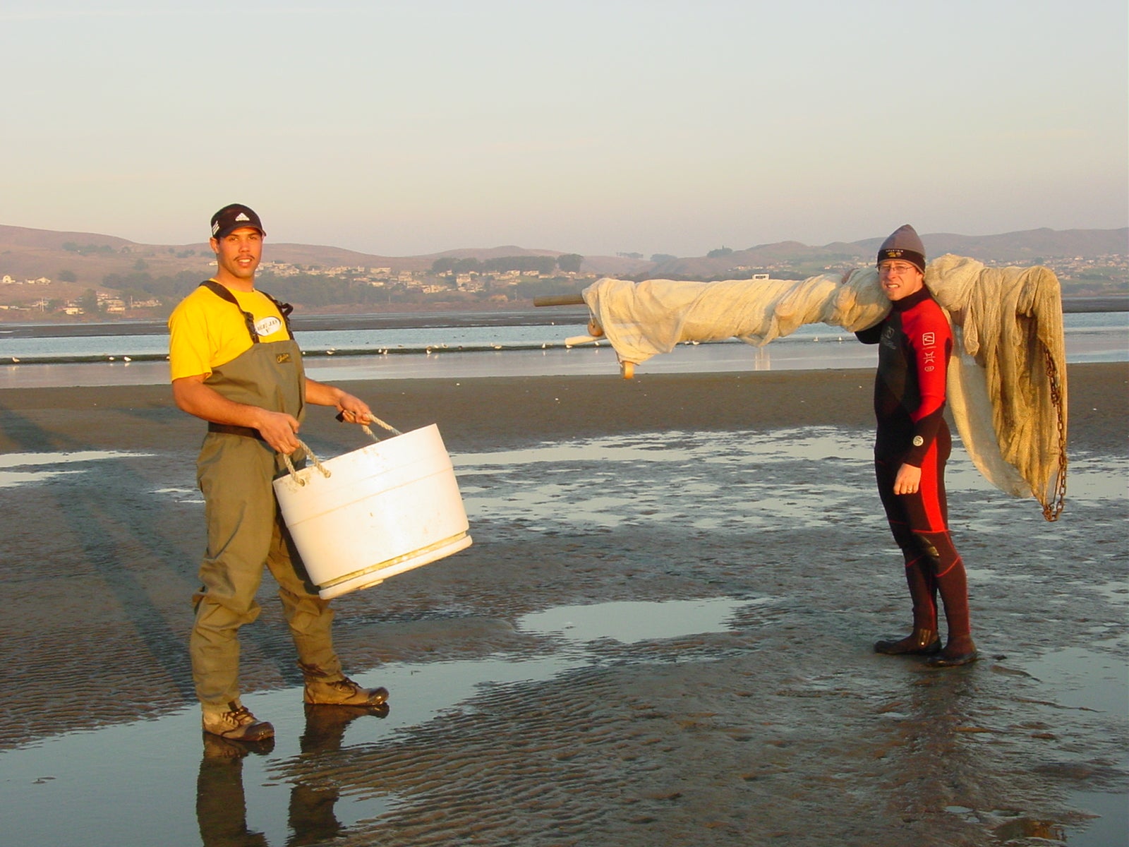 man holding white tub and man in red and black wetsuit on tidal marsh