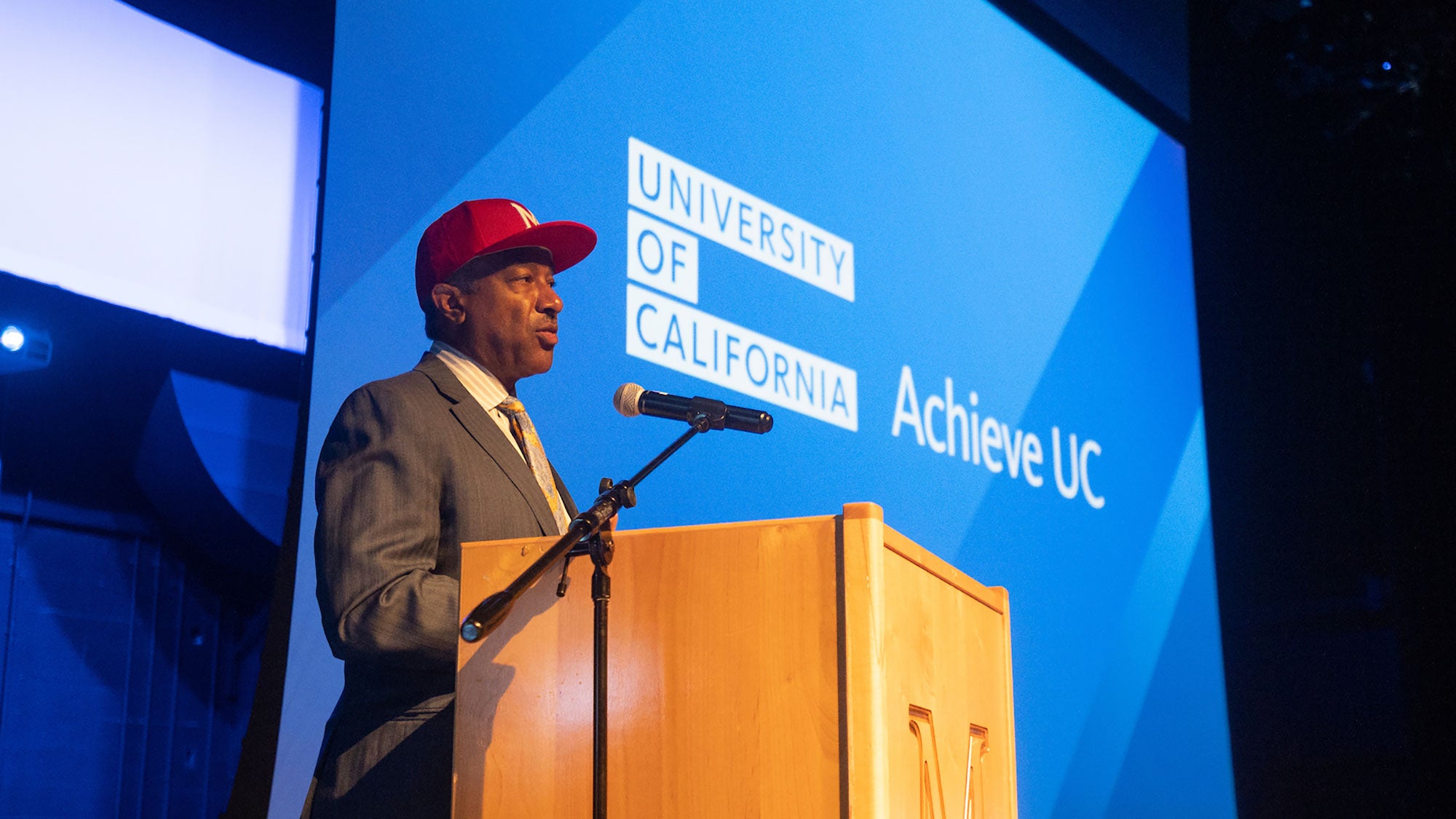 Chancellor Gary S. May wearing a baseball cap and standing on stage at a lectern