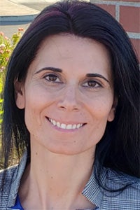 Smiling woman with dark hair against brick wall and sunlit greenery