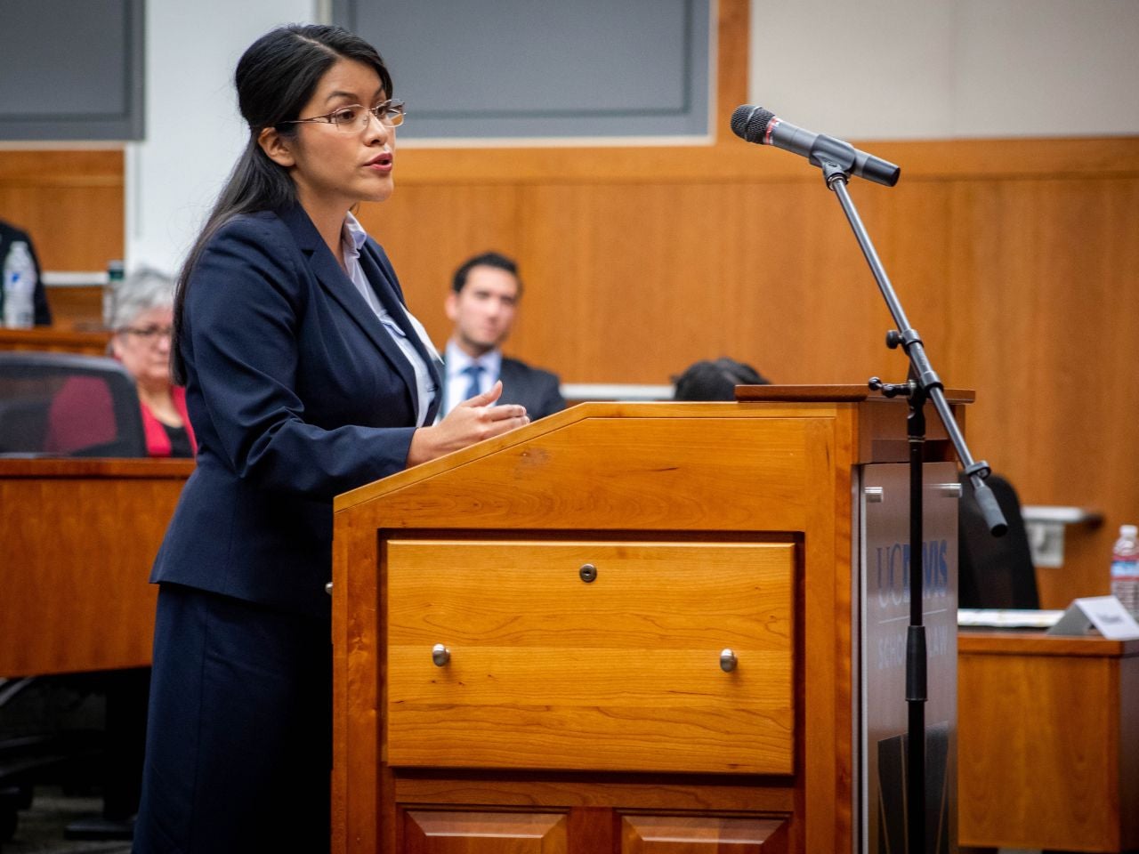 A student in business attire stands at a podium, speaking into a microphone, while onlookers listen.